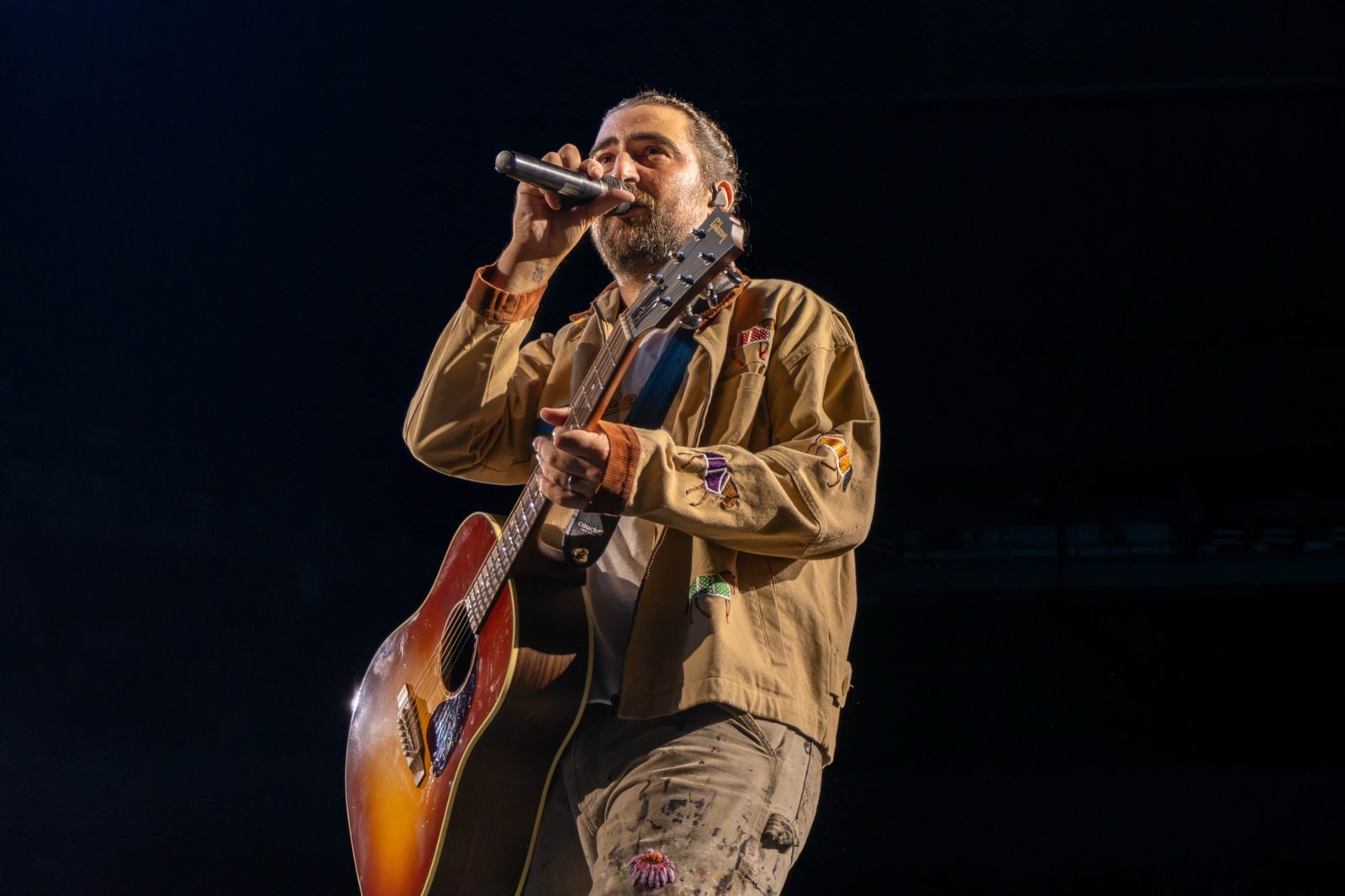 Noah Kahan singing into the mic with his acoustic guitar, black background