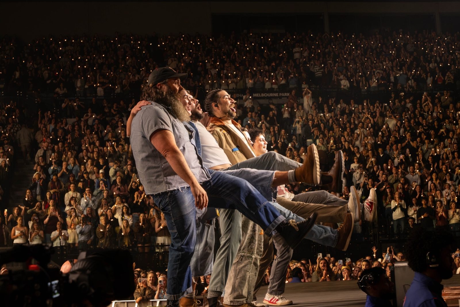 Band members on stage edge, one kicking up a leg, arena crowd behind